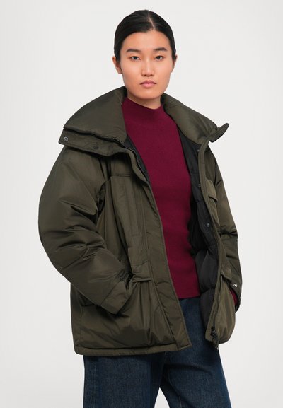 Woman wearing an oversized olive green puffer jacket over a burgundy sweater and blue jeans, standing against a plain white background.