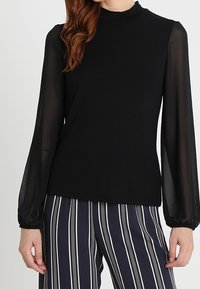 Svart topp med hög hals, långa genomskinliga ärmar och tight passform, tillsammans med randiga vida byxor. Mjuk textur, minimalistisk design.