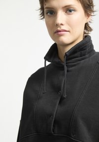 Pullover in cotone nero con colletto alto, dettagli con bottoni e cordini. Presenta una texture morbida e cuciture tonali.