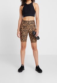 Černá sportovní podprsenka a cyklistické šortky s leopardím potiskem. Látka je hladká a elastická. Modelka drží černou láhev na vodu a má na sobě černé tenisky.