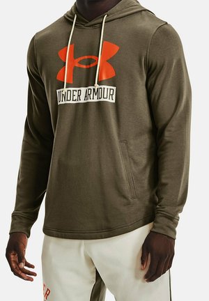 Man draagt een olijfgroene Under Armour hoodie met oranje logo en crèmekleurige broek met rode letters op een witte achtergrond.