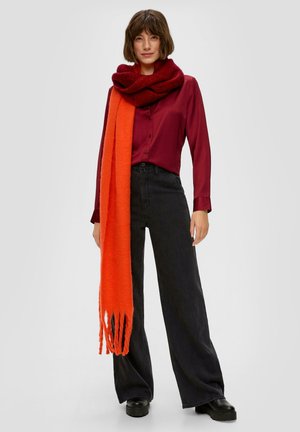 Rode en oranje gelaagde sjaals van zachte stof; model draagt een bordeaux blouse en wijde zwarte broek; casual styling met een ontspannen pasvorm.