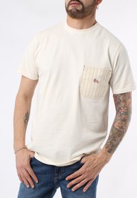 T-shirt in cotone color crema con collo rotondo, caratterizzato da una tasca a righe con logo rosso. La vestibilità è rilassata, mettendo in mostra i tatuaggi su un braccio.