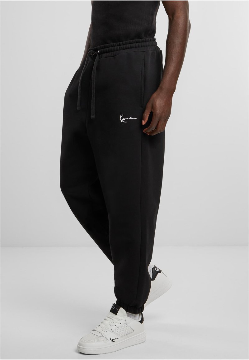 Schwarze Sweatpants mit lockerer Passform, ausgestattet mit einem elastischen Bund, Kordelzug und einem kleinen weißen Logo auf dem Oberschenkel. Kombiniert mit weißen Sneakers.