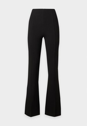Frau steht, trägt ein schwarzes ärmelloses Top mit quadratischem Ausschnitt und passende hoch taillierte Hosen mit vorderen Schlitzen sowie schwarze Loafers.