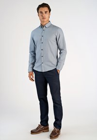 Lichtblauw overhemd met lange mouwen, donkere knopen, gecombineerd met donkere marineblauwe broeken en bruine bootschoenen op een neutrale achtergrond.