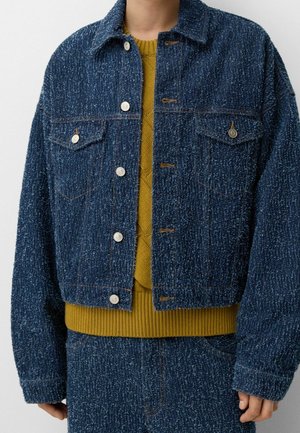 Person iført en tekstureret mørkeblå jakke med knapper over en sennepsgul strikket sweater og matchende mørkeblå bukser.