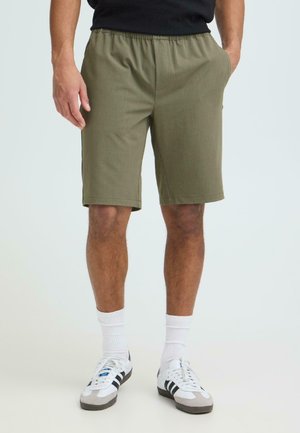 Mand iført olivengrønne shorts til knæet, en sort skjorte, hvide mellemlange sokker og hvide sneakers med sorte striber, stående med hænderne i lommerne.