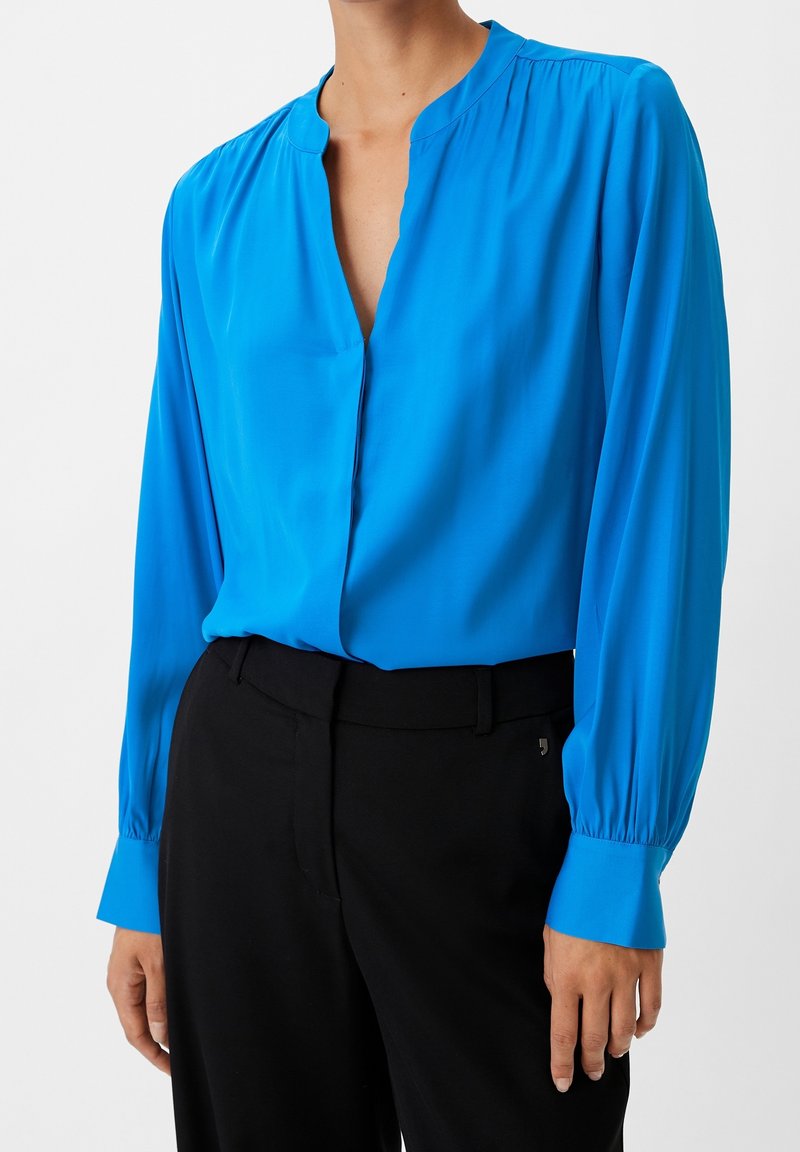 Blusa azzurra brillante con maniche lunghe, scollo a V profondo e spalle arricciate. Indossata con pantaloni neri, caratterizzati da polsini aderenti. Tessuto morbido.