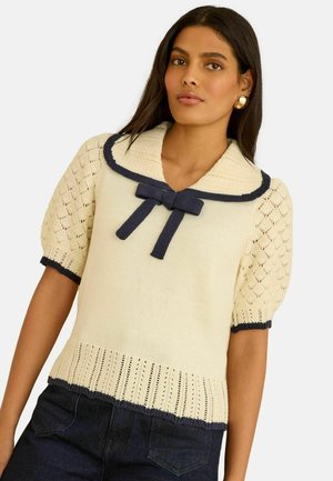 Frau mit langen dunklen Haaren, die einen cremefarbenen, kurzärmeligen Strickpullover mit navyblauem Kragenrand und Schleife trägt, kombiniert mit dunkler Jeanshose.