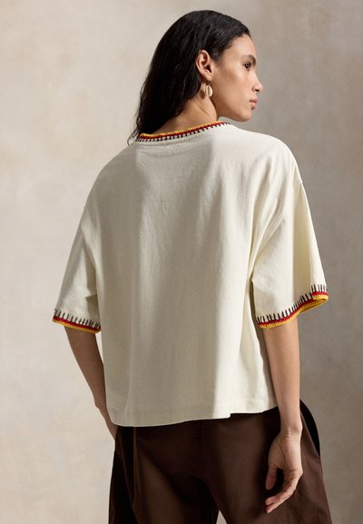 Woman with dark hair wears oversized cream shirt with red and yellow stitched trim on sleeves and neckline, paired with dark brown pants.