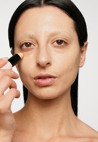 Bâton de maquillage à la main, appliquant une crème dans une teinte pêche claire sous l'œil. Texture de peau lisse ; cheveux foncés et piercing visibles.