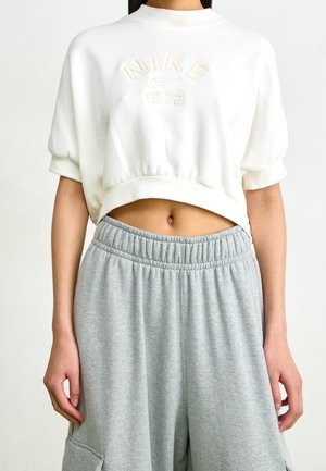 Frau trägt weißes, kurzes Nike Sweatshirt mit dem Text "1972" und graue Jogginghose mit elastischem Bund, zu sehen vom Hals bis zur Hüfte.