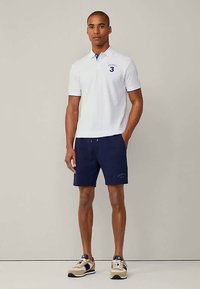 Άνδρας που στέκεται φορώντας λευκό πόλο μπλουζάκι με τον αριθμό 3, ναυτικό μπλε σορτς και μπεζ αθλητικά παπούτσια, μπροστά από λευκό φόντο.