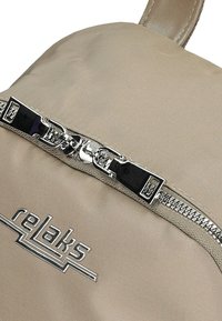 Beżowy plecak z błyszczącym, srebrnym zamkiem błyskawicznym z czarnym akcentem oraz logo "relaks" w metalicznie srebrnym haftem z przodu.