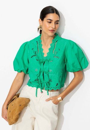 Femme portant un chemisier vert brodé avec des manches bouffantes et un pantalon crème, tenant une pochette en daim beige, accessoirisée avec des boucles d’oreilles et un bracelet dorés.