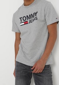 Gray cotton t-shirt with "TOMMY JEANS" in bold navy letters and a red and white logo accent, round neck, and short sleeves.