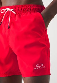 Pantaloni da bagno rossi realizzati in tessuto liscio, con vita elastica e cordino bianco, e un logo sul lato destro in basso.