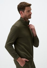 Olive green pulower z zamkiem ćwierć, wykonany z teksturowanej tkaniny, z długimi rękawami, ściągaczami oraz luźnym krojem dla komfortu.