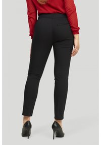 Pantalon noir ajusté avec une texture lisse, jambes fuselées et une poche arrière. Porté avec des talons hauts noirs. Chemisier rouge visible en haut.
