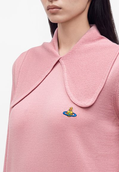 Femme portant un haut en maille rose avec un grand col et un petit logo brodé coloré sur la poitrine.