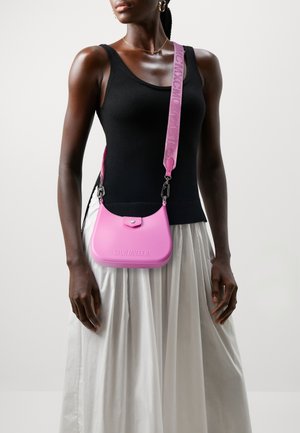 Femme portant un haut sans manches noir et une jupe blanche, avec un sac bandoulière rose Steve Madden à large bandoulière sur l'épaule.