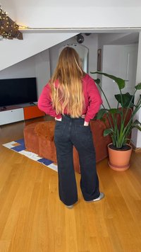 Μαύρο τζιν παντελόνι υψηλής μέσης με φαρδιά μπατζάκια και δύο πίσω τσέπες, συνδυασμένο με κόκκινο μακρυμάνικο τοπ. Εσωτερικός χώρος με φυτά και έπιπλα ορατά.