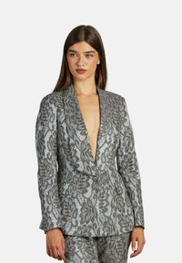 Blazer argento con scollatura a V profonda, caratterizzato da un intricato motivo floreale, texture liscia e vestibilità sartoriale. Pantaloni coordinati visibili.