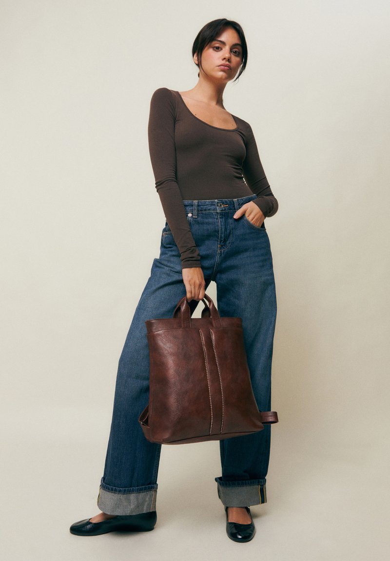 Femme portant un haut à manches longues foncé et un jean large tient un grand sac cabas en cuir marron, debout devant un fond clair uni.