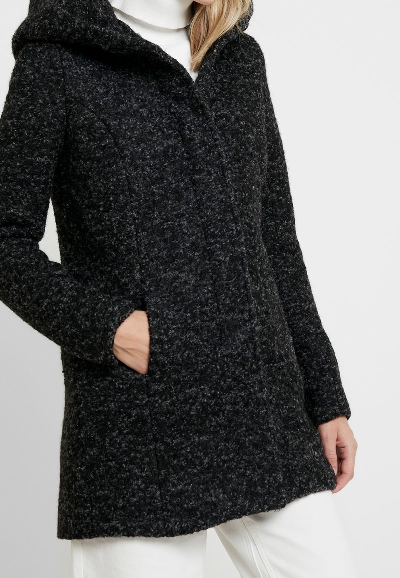 Frau trägt einen strukturierten dunkelgrauen Kapuzenmantel mit Seitentaschen über einem weißen Rollkragenpullover und weißen Hosen.