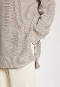 Light beige knit sweater with ribbed texture, wide sleeves, and side slits. Paired with cream-colored corduroy pants.