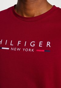 Rotes Baumwoll-T-Shirt mit Rundhalsausschnitt und weißem Text mit "HILFIGER" und "NEW YORK", akzentuiert durch horizontale Streifen in Marineblau und Rot.