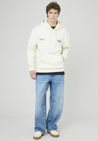 Sweatshirt crème avec des détails textuels à l'avant, associé à un jean bleu clair ample et des baskets beiges. Tissu doux, design décontracté et coupe décontractée.