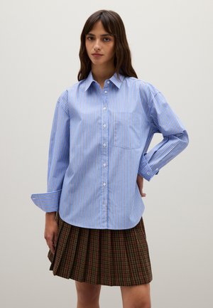 Camisa de botones a rayas azul y blanca con mangas largas, un bolsillo en el pecho izquierdo y un corte relajado. Combinada con una falda plisada de textura oscura.