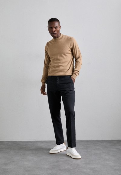 Beige stickad tröja, svarta skräddarsydda byxor och vita sneakers med bruna detaljer. Modellen står mot en neutral bakgrund.