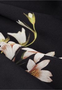 Černá tkanina s bílými květinami a zelenými akcenty, s propracovanými detaily a jemnou texturou. Vzor zdůrazňuje květinový design.