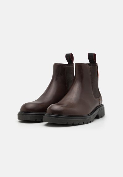 Brown leather Chelsea boots with elastic side panels, rounded toe, textured upper, and a durable black rubber sole. Red accent on the heel.