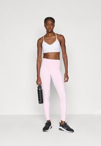 Fehér sportmelltartó, rózsaszín leggings és fekete sportcipő. Fekete vízpalackot tart a kezében. Simább anyag, testhezálló dizájn, minimális márkajelzés.