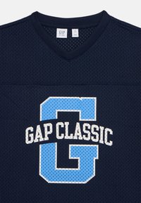 Jūras spēku tīkla džersijs ar V veida kaklu, ar baltu un gaiši zilu "GAP CLASSIC" tekstu un lielu kontūrveida "G" grafiku. Standarta piegriezums.