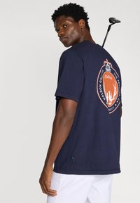 Námornícky modré bavlnené tričko s veľkým oranžovým logom na chrbte, s okrúhlym strihom a krátkymi rukávmi. Obsahuje zelený akcent na leme.