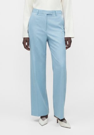 Personne portant un pantalon large bleu clair sur mesure, un haut blanc à manches longues et des talons hauts blancs pointus avec des détails en dentelle.