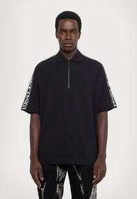 Mladý muž v čiernom polo tričku s krátkym rukávom, na ktorom sú pruhy so sloganom "Versace Jeans Couture", a v čiernych nohaviciach posypaných bielym farbom.