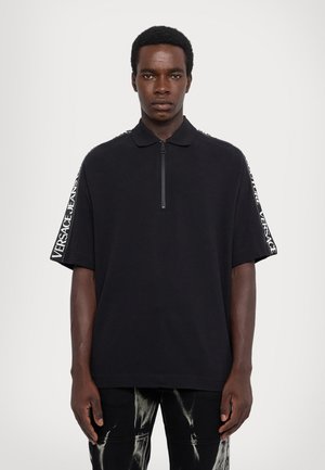 Ung mann iført en svart kortermet polo med "Versace Jeans Couture" tekst på ermelinjer og svarte bukser med hvite malingsplatter.