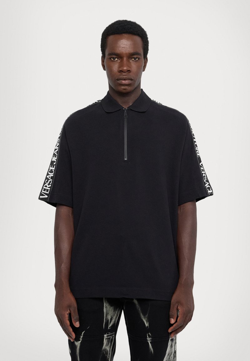 Mladý muž v čiernom polo tričku s krátkym rukávom, na ktorom sú pruhy so sloganom "Versace Jeans Couture", a v čiernych nohaviciach posypaných bielym farbom.