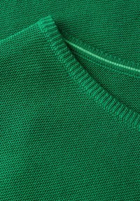 Grön stickad tröja med texturerad yta och ribbad halsringning. Materialet verkar mjukt och av medelvikt.