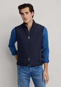 Mladý muž v tmavě modré prošívané vestě na zip přes modrou košili s vyhrnutými rukávy a modré džíny, stojící před jednobarevným pozadím.