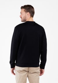 Hombre con cabello corto castaño, vistiendo un sudadera negra y pantalones beige, girado para mostrar la vista trasera sobre un fondo blanco.