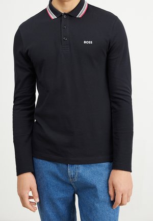 Mężczyzna w czarnej, długorękawowej koszulce polo z prążkowanym kołnierzykiem i białym logo BOSS, zestawionej z niebieskimi jeansami.