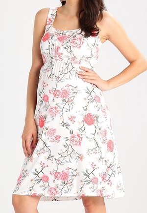 Witte mouwloze jurk met roze bloemenprint, gerimpelde halslijn en een uitlopende rok tot op de knie, gedragen door een staande vrouw met hand op haar heup.