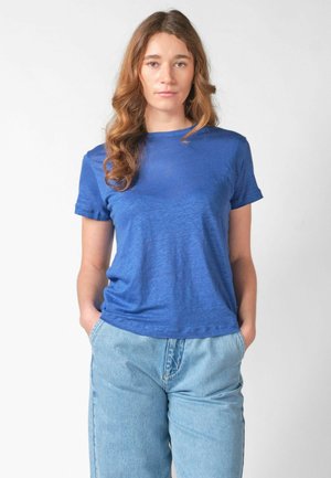 Tânără cu păr castaniu ondulat, purtând un tricou albastru și blugi deschiși la culoare, stând cu mâinile în buzunare pe un fundal simplu.